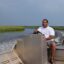 He is seated behind the wheel of a metal boat, the river bending behind him.